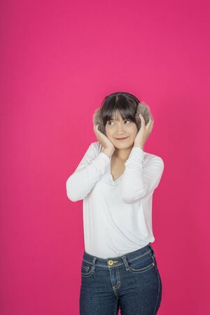 Adorable asian woman in white shirt singing and having fun while listening to music using wireless earphones isolated over pink backgroundの写真素材