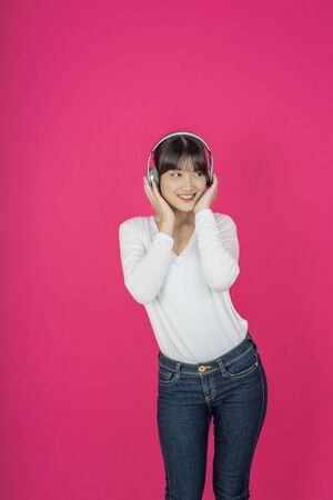 Adorable asian woman in white shirt singing and having fun while listening to music using wireless earphones isolated over pink backgroundの写真素材