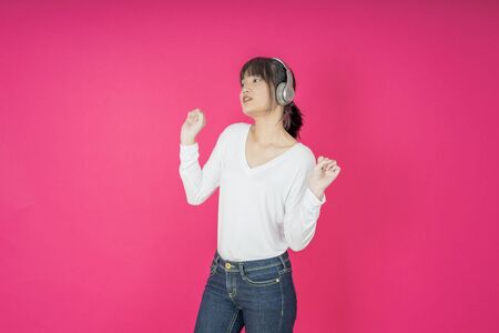 Adorable asian woman in white shirt singing and having fun while listening to music using wireless earphones isolated over pink backgroundの写真素材