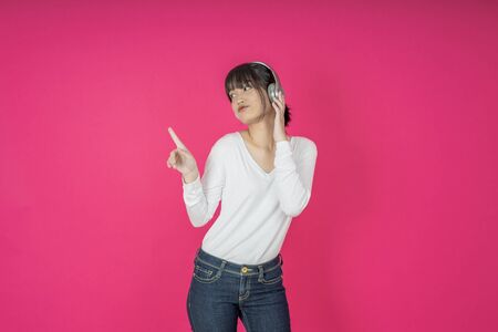 Adorable asian woman in white shirt singing and having fun while listening to music using wireless earphones isolated over pink backgroundの写真素材