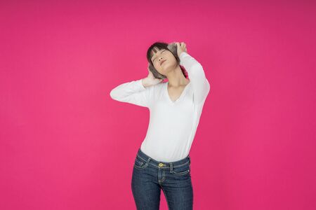 Adorable asian woman in white shirt singing and having fun while listening to music using wireless earphones isolated over pink backgroundの写真素材