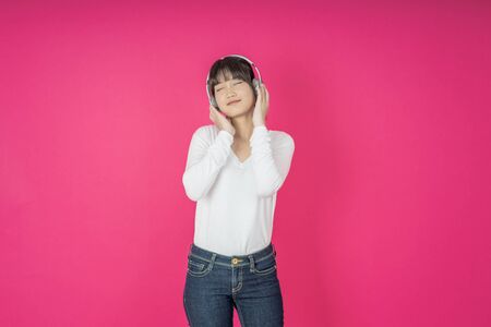 Adorable asian woman in white shirt singing and having fun while listening to music using wireless earphones isolated over pink backgroundの写真素材