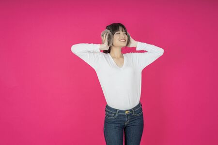Adorable asian woman in white shirt singing and having fun while listening to music using wireless earphones isolated over pink backgroundの写真素材