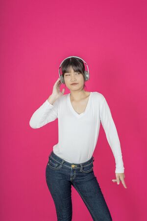 Adorable asian woman in white shirt singing and having fun while listening to music using wireless earphones isolated over pink backgroundの写真素材