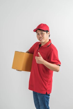 Young smiling logistic delivery man in red uniform holding the box on white backgroundの写真素材