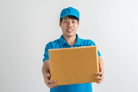 Young smiling logistic delivery man in blue uniform holding the box on white background. Asian peopleの写真素材