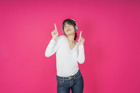Adorable asian woman in white shirt singing and having fun while listening to music using wireless earphones isolated over pink backgroundの写真素材