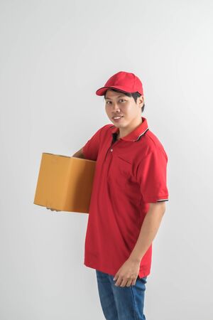 Young smiling logistic delivery man in red uniform holding the box on white backgroundの写真素材