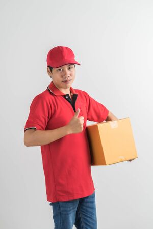 Young smiling logistic delivery man in red uniform holding the box on white backgroundの写真素材