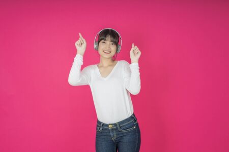 Adorable asian woman in white shirt singing and having fun while listening to music using wireless earphones isolated over pink backgroundの写真素材