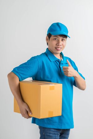 Young smiling logistic delivery man in blue uniform holding the box on white background. Asian peopleの写真素材