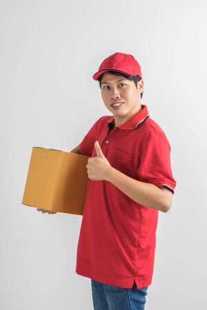 Young smiling logistic delivery man in red uniform holding the box on white backgroundの写真素材