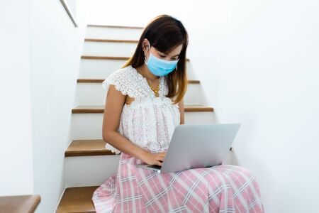 Asian woman working or learn from home with laptop and medicine health care mask on stairの写真素材