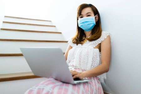 Asian woman working or learn from home with laptop and medicine health care mask on stairの写真素材
