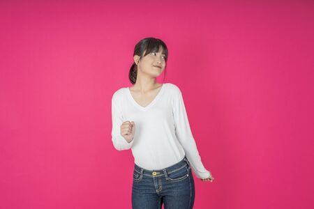 Adorable asian woman in white shirt singing and having fun while listening to music using wireless earphones isolated over pink backgroundの写真素材