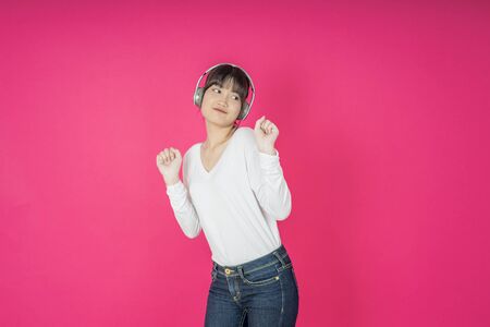 Adorable asian woman in white shirt singing and having fun while listening to music using wireless earphones isolated over pink backgroundの写真素材