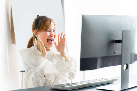 Portrait of happy young asian woman celebrating success with arms up in front of laptop at home.