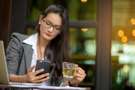 Young female freelancer checking email on cell during tea break with bokeh background. Business concept.の写真素材