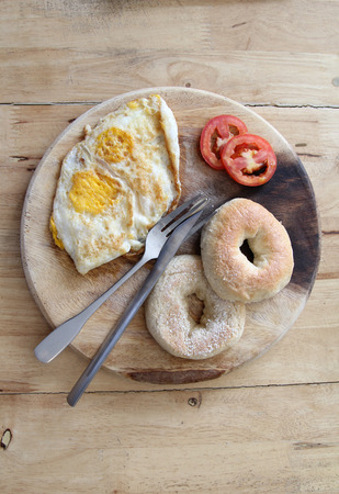 Breakfast; donut, fried egg and tomato with knife and fork on wooden plate.の写真素材