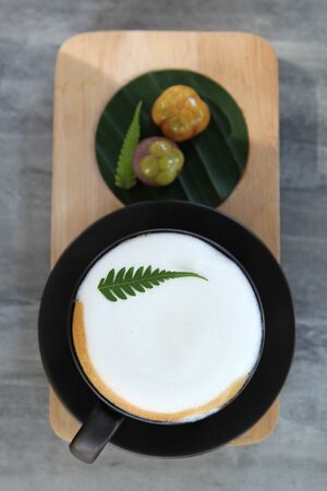Hot cappuccino decorated by fern leaf in black cup with Thai  dessert on wooden plate.の写真素材