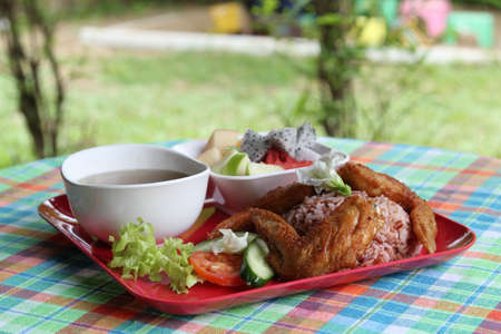 Thai style food, the deep fried chicken wings with rice, soup, vegetable and fruit on red plate.の写真素材