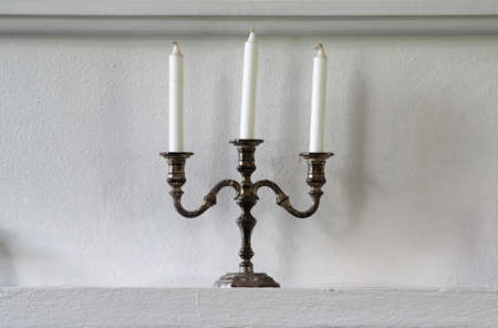 Antique candlestick with three white candles on white wall.