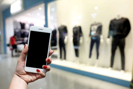 woman holds a smartphone standing in front of a mall. There is a blurred image of the clothing store as the background.の写真素材