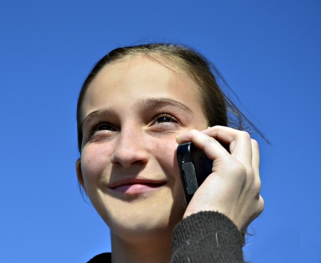 Beautiful preteen girl talking on a cell phone with blue sky background.の写真素材