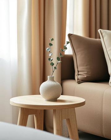 A serene interior shot featuring a small, round wooden side table. A textured vase holds delicate eucalyptus branches, set against a backdrop of soft curtains and a neutral-toned couch.の素材