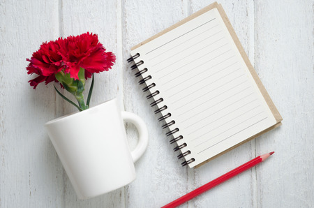 Empty notepad, pencil and red Carnation flowers in a cup  on the white wooden background, top view.の写真素材