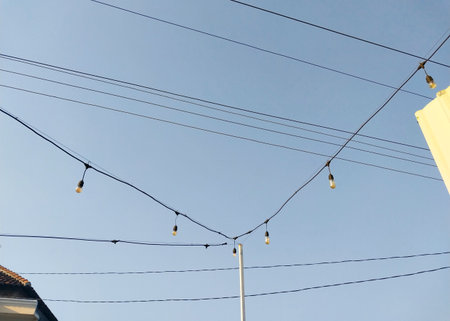 Outdoor string lights hanging across utility poles against a clear blue sky in the afternoon, creating a minimal, serene, and urban aesthetic with visible power lines and rooftopsの写真素材