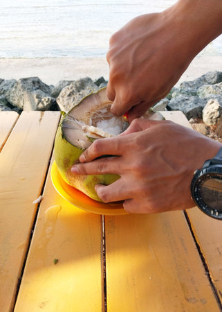 A close-up of hands skillfully cutting open a fresh coconut on a sunny beachside table, revealing...