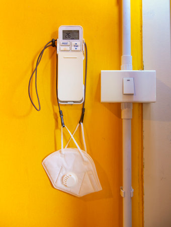 A medical mask or hygienic mask hangs on an air purifier remote control mounted on an orange wall