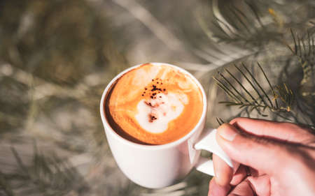A man's hand holds a cup of white cappuccino filled with cream and sprinkled with cinnamon.warm and vintage photographyの写真素材
