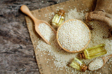 Top view of dry organic white sesame seeds in wooden bowl over blur background of some on sack fabric and grunge floor with sesame oil in glass bottle, on dark brown toneの写真素材