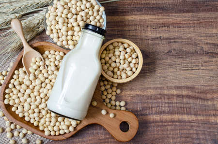 Top view of dry organic homemade soybean milk in glass bottle with some seeds in wooden bowl on sack fabric with dry wheat ear on dark brown wooden backgroundの写真素材
