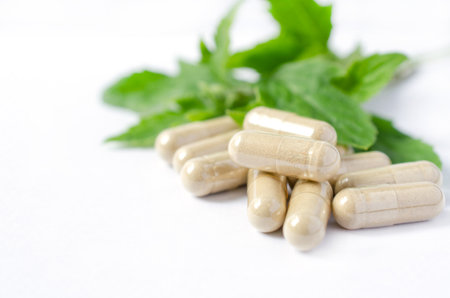Closed up green Thai herb powder in capsule on blur green herb leaf on white background. For natural medication or alternative choice of disease treatment to avoid chemical allergyの写真素材