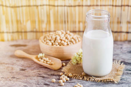 Closed up fresh organic homemade soybean milk in glass bottle over blur soybean seed pile in wooden bowl and spoon on grunge background in soft light toneの写真素材