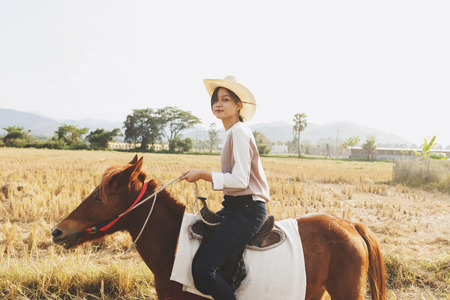 Beautiful young woman is smiling riding a horse on the field with relax time . Sideways to the camera. Freedom, joy, movementの写真素材