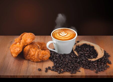 Hot coffee, coffee beans and croissants rest on an old wooden table with a black and red backdrop.の写真素材