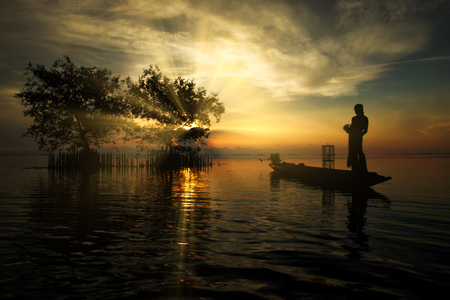 Silhouettes fisherman on the fishing boat and tree in the lake with sunrise.の写真素材