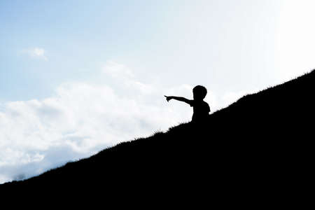 Silhouette of a boy pointing forward on the hill with sky background.の写真素材