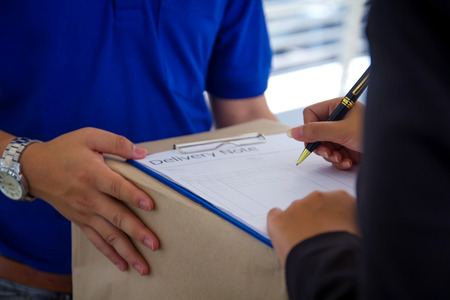 Close up Delivery man in blue uniform holding package while woman is signing  receiving documents.Delivery man delivering parcel boxes to a woman customer.の写真素材