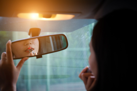 Young woman looking in rear view mirror and putting make up in car. modern busy life.beautiful girl sitting in drivers seat of car doing make up, applying lipstick.の写真素材