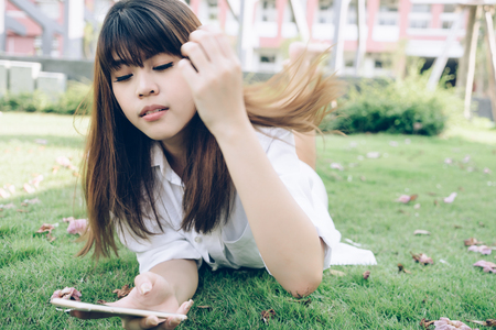 Young woman lying on green grass floor holding mobile phone with hair waving. Portrait fashion model girl in outdoor nature.の写真素材