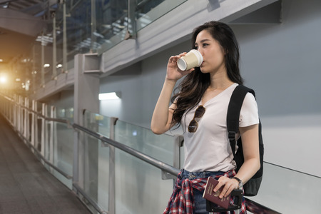 Beautiful young Asian woman arrives airport early morning ready going aboard foreign country.Tourist girl with backpack carryon luggage in airport terminal.Female traveler drinking coffee in international airport holding Thai passport and suitcase or baggage.の写真素材