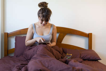 Young Asian woman using modern digital tablet sitting on bed making payment for purchase in web...