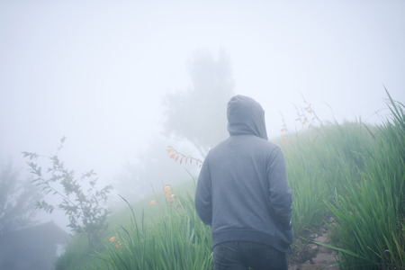 Hipster man in hood walking through spooky foggy winters day, back view.