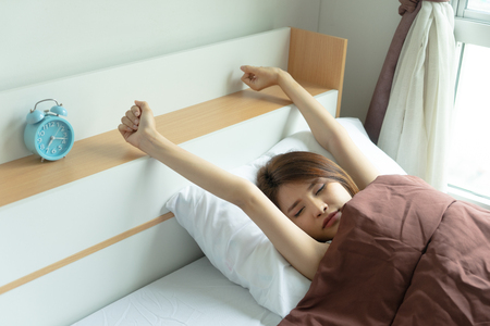Young woman stretch arms in the air with alarm clock. wake up early in morning. Girl stop snooze alarm. female waking up sleepy and tired. woman wake up in the morning with alarm clock snooze.の写真素材