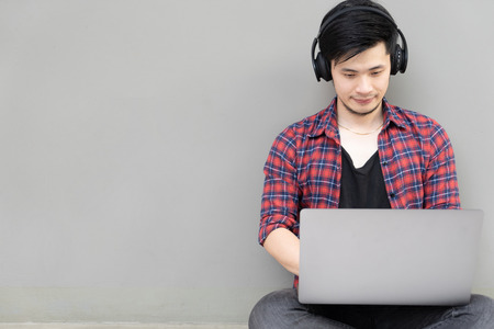 Young Asian businessman sitting on floor working using laptop computer while listening to music with headphone against gray background.
Music network streaming relaxation technology concept.
Focused man studying with laptop, concentrated student or office worker listening to audio business course at work, e-learning and online professional education concept.の写真素材